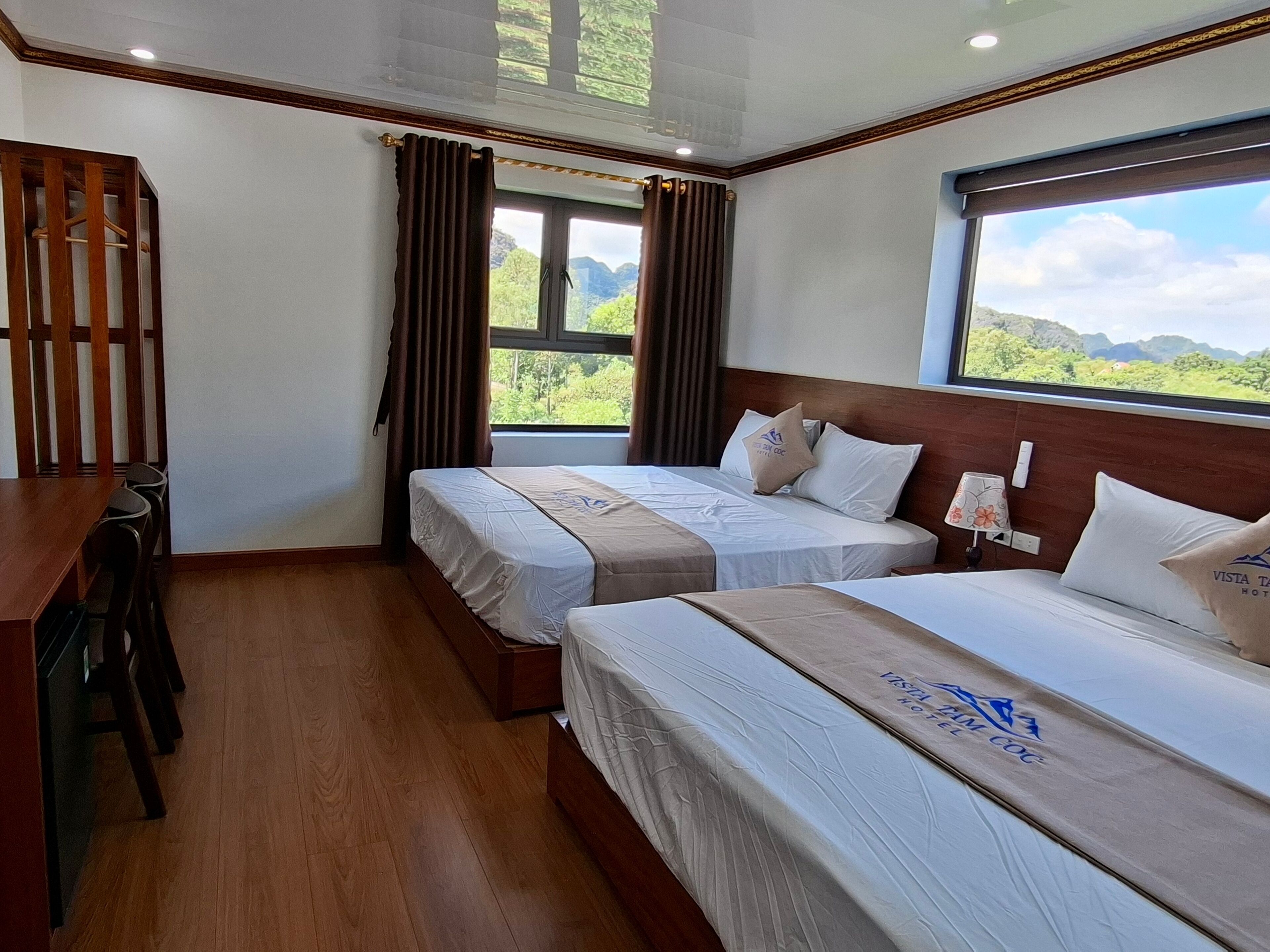 A hotel room with two beds, a wooden desk, and a window with a view of greenery.