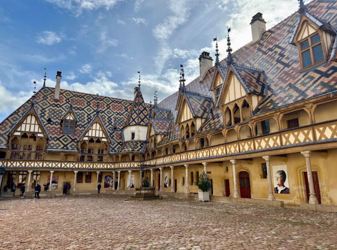 A courtyard with a cobblestone floor, a building with a patterned roof, and a statue in the center.