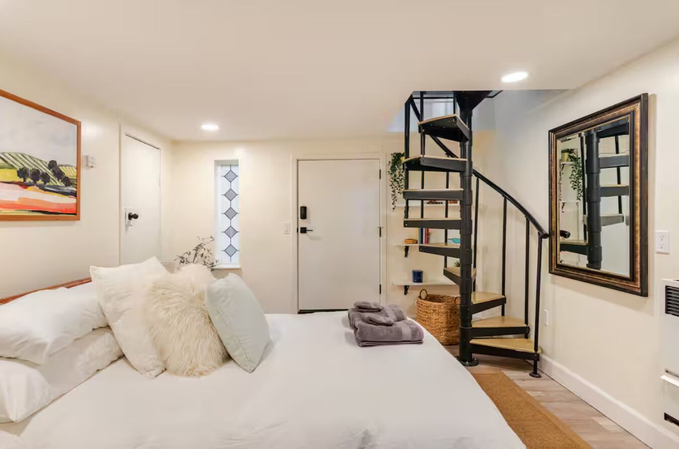 A bedroom with a bed, a white door, a black spiral staircase, and a framed mirror on the wall.