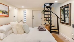 Un dormitorio con cama, una puerta blanca, una escalera en espiral negra y un espejo enmarcados en la pared.