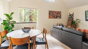Una sala de estar moderna con una mesa de comedor redonda, sillas de madera, un sofá gris y un ventanal con vistas a árboles.