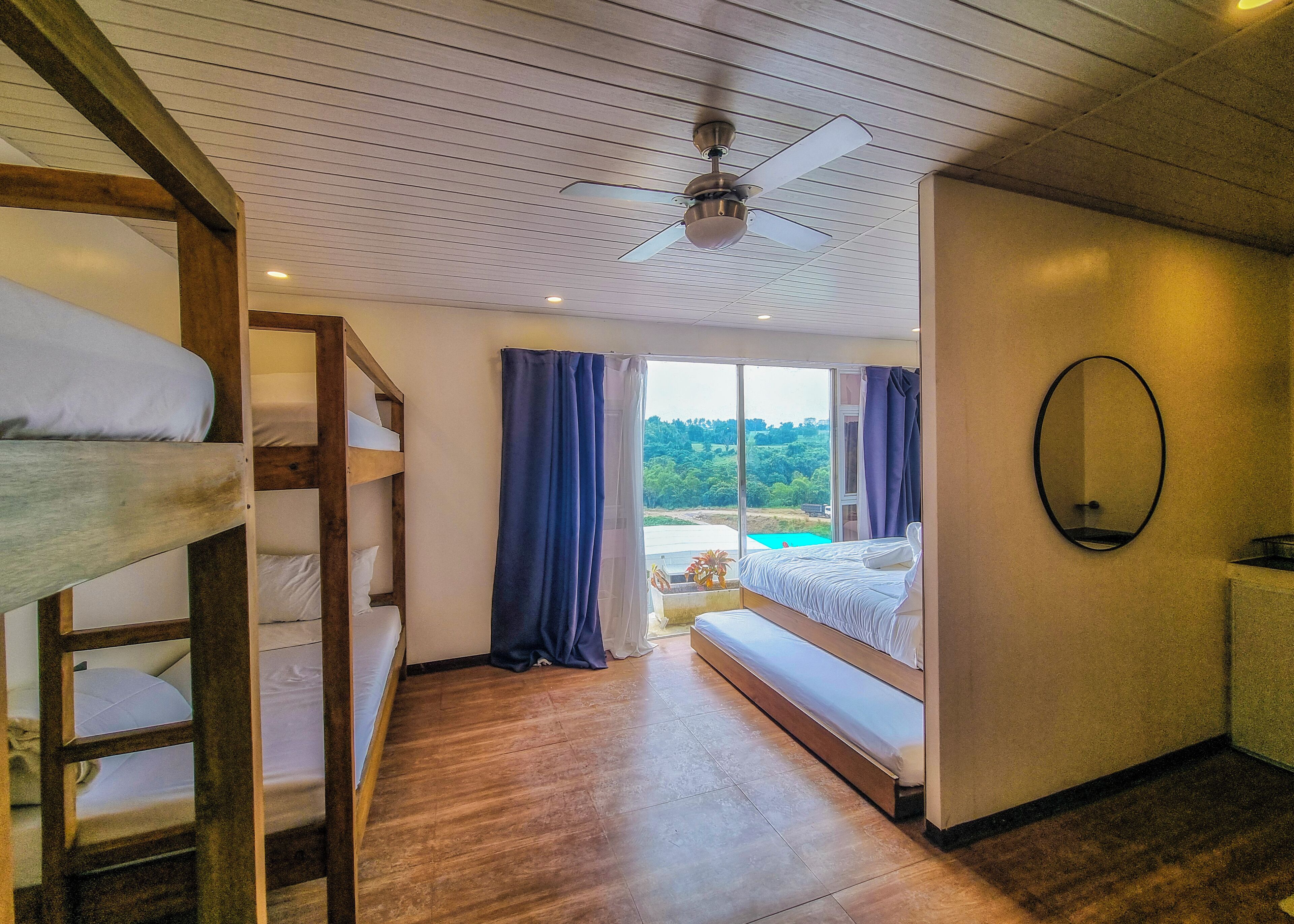 A room with bunk beds, a ceiling fan, a large window with curtains, and a view of greenery outside.
