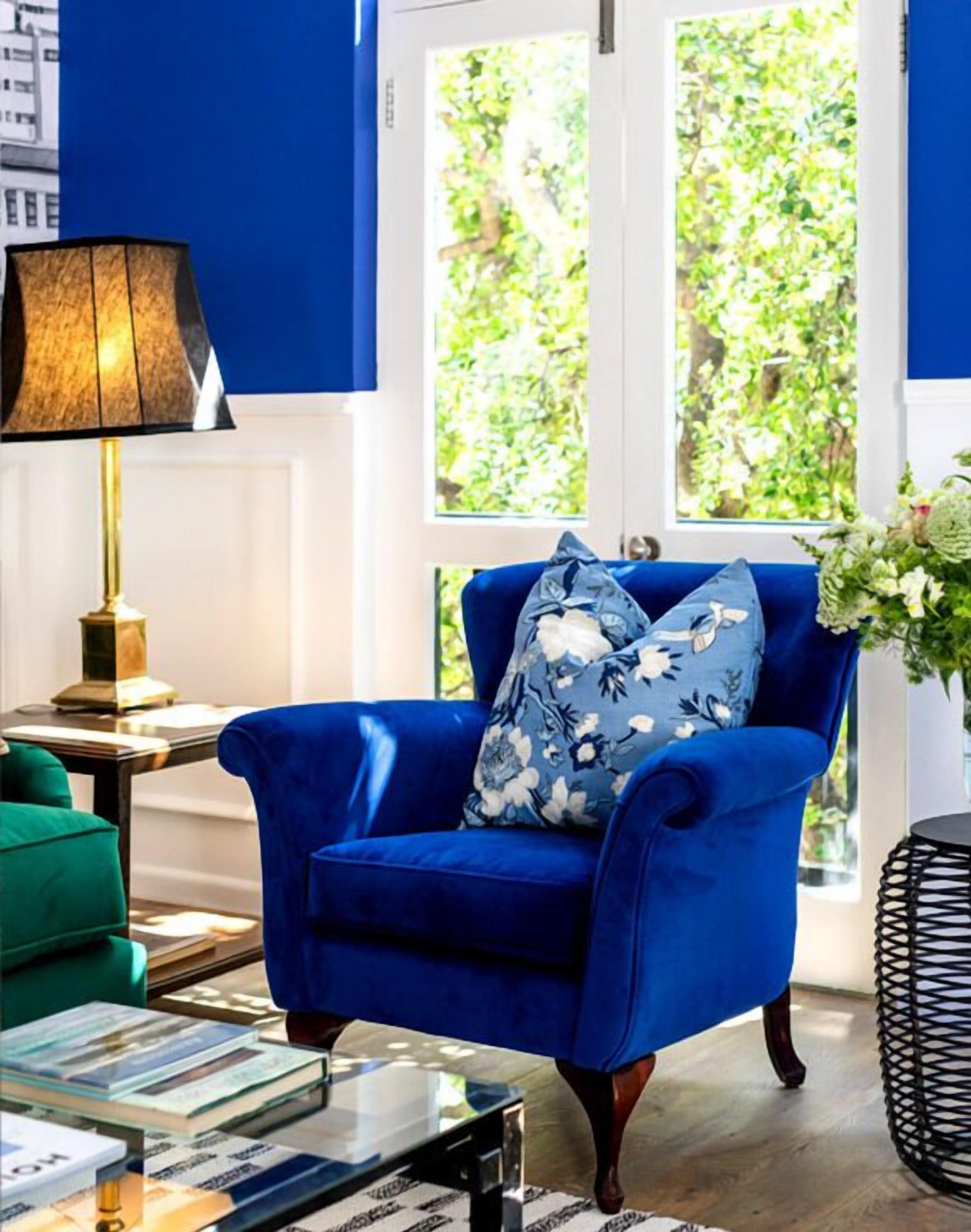 Un sillón azul con almohadones florales, un sofá verde, una lámpara y un jarrón con flores.