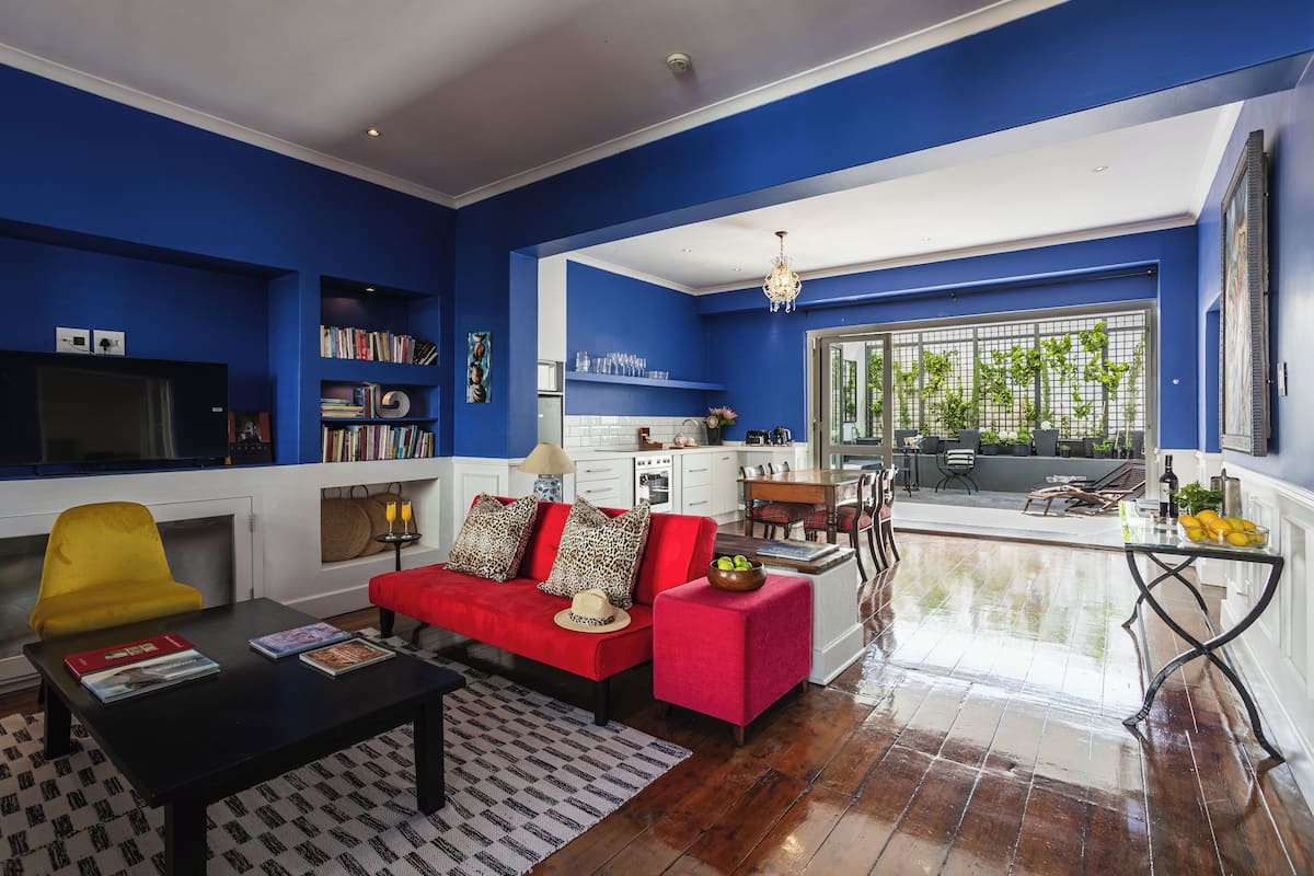 A living room with a red sofa, a yellow chair, and a black coffee table. There is a bookshelf with books and a TV on the left. The room has blue walls and a large window with plants.