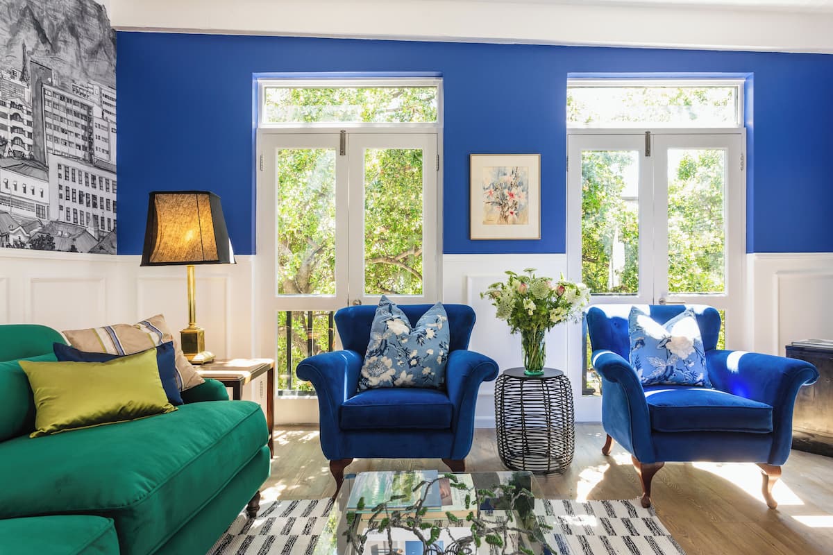 A room with a blue wall, two blue armchairs with floral cushions, a green sofa, a glass coffee table, and a black side table with a lamp.