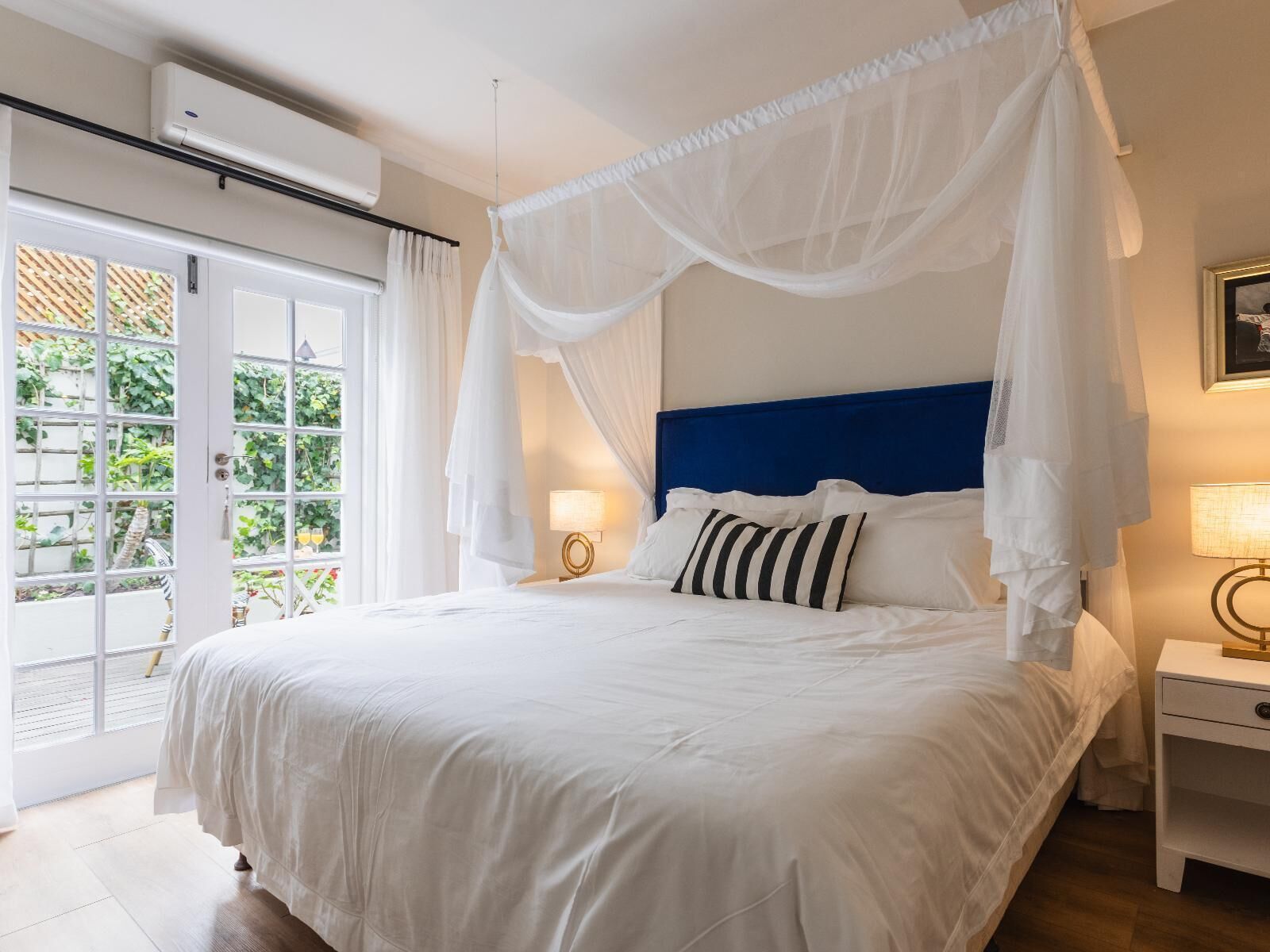 A bedroom with a canopy bed, a bedside table, and a view of greenery through a glass door.