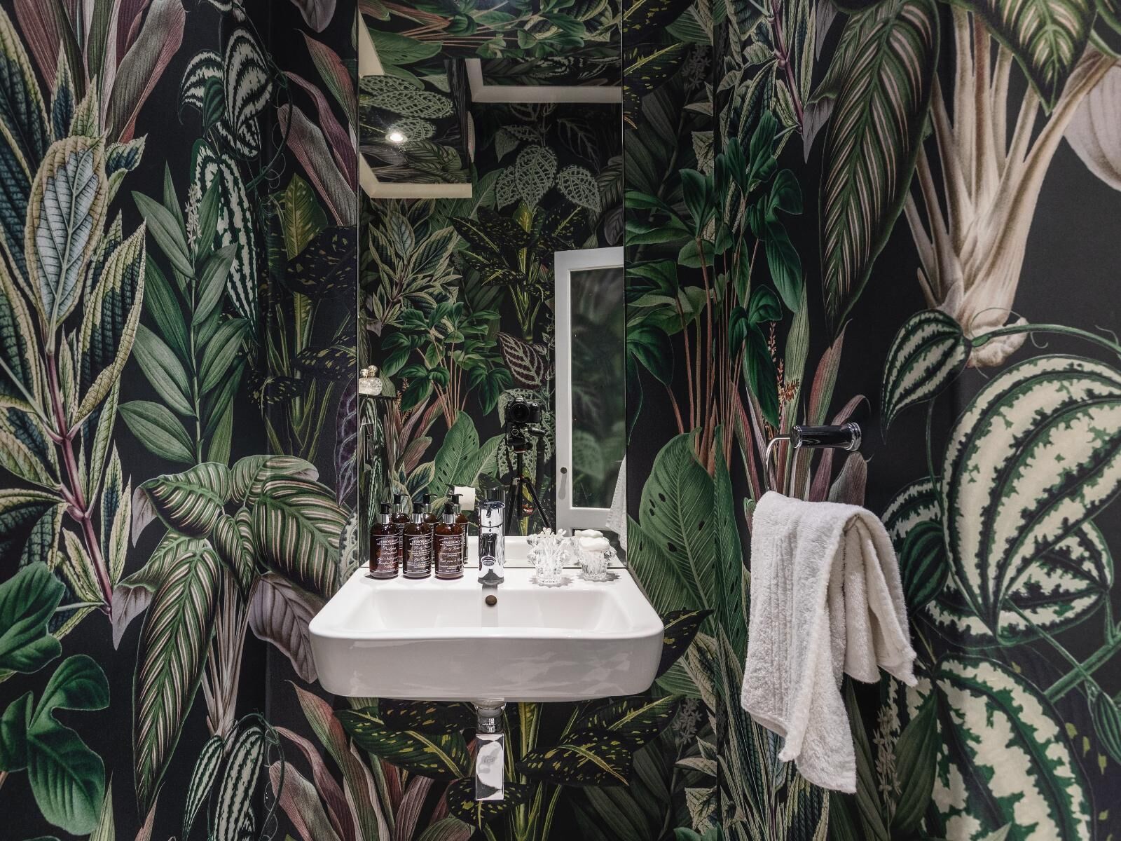 A bathroom with a white sink, a mirror, and a towel rack. The walls are decorated with a tropical leaf pattern.