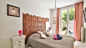 A bedroom with a wooden headboard, a white bedside table with a lamp, a bed with pillows and a rolled towel, and a view through a glass door.
