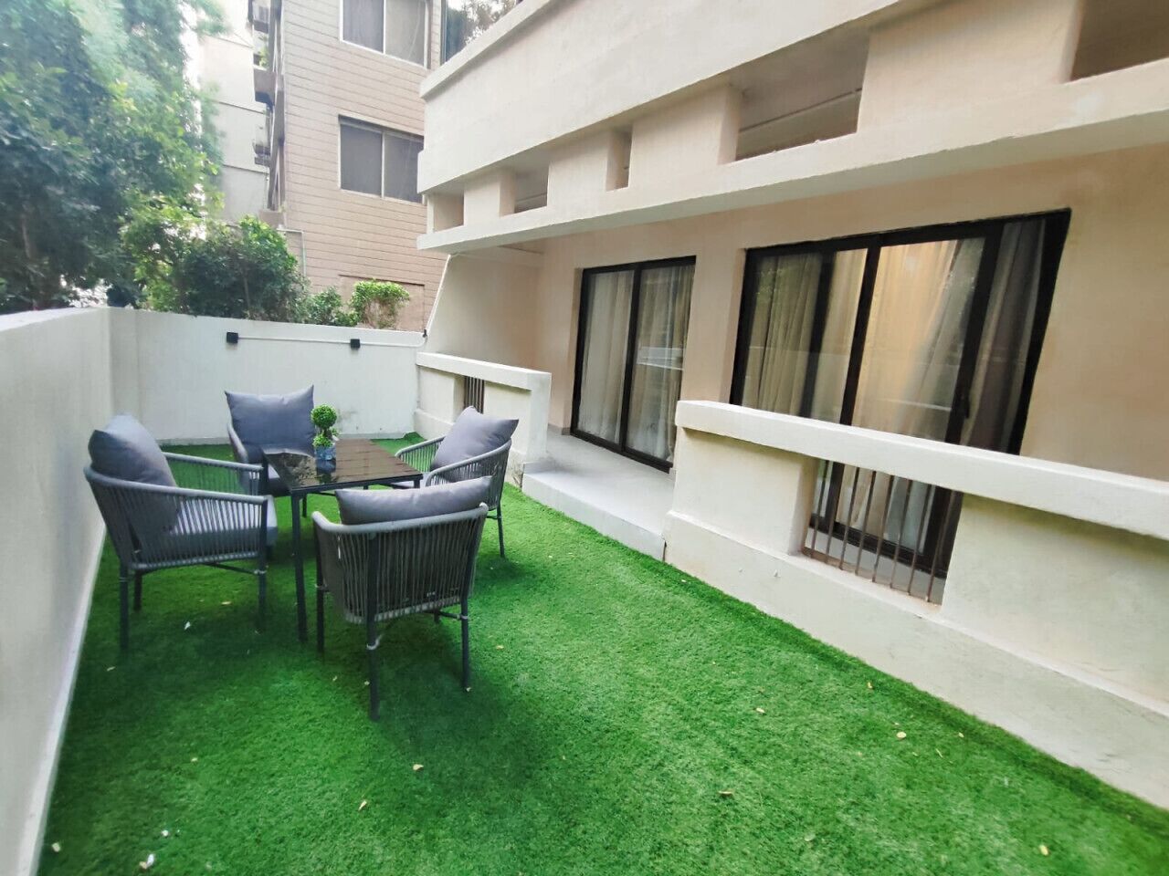 Un pequeño patio al aire libre con una mesa y sillas, rodeado por un muro blanco y un edificio con ventanas amplias.