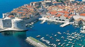 Vista aérea de uma cidade costeira histórica, com um castelo fortificado, ruas estreitas e uma marina repleta de barcos.