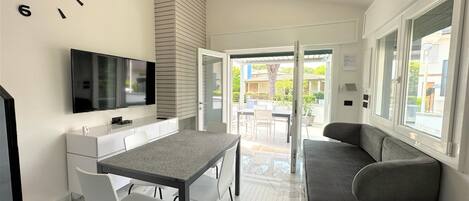 A modern kitchen with a flat-screen TV, a dining table with chairs, and a sofa by a large window.