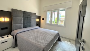 Un dormitorio moderno con una cama grande, una mesita de noche, una lámpara de pared y una ventana que ofrece vistas a edificios.