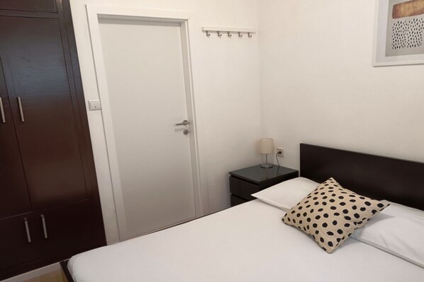 A bedroom with a bed, a nightstand, a lamp, a polka dot pillow, a wardrobe, and a door. - Two Bedroom Apartment (Povlja)