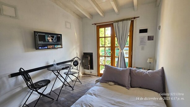 A room with a bed, a TV mounted on the wall, a small table with chairs, a window with curtains, and a door.