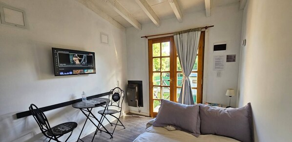 A room with a bed, a TV mounted on the wall, a small table with chairs, a window with curtains, and a door.