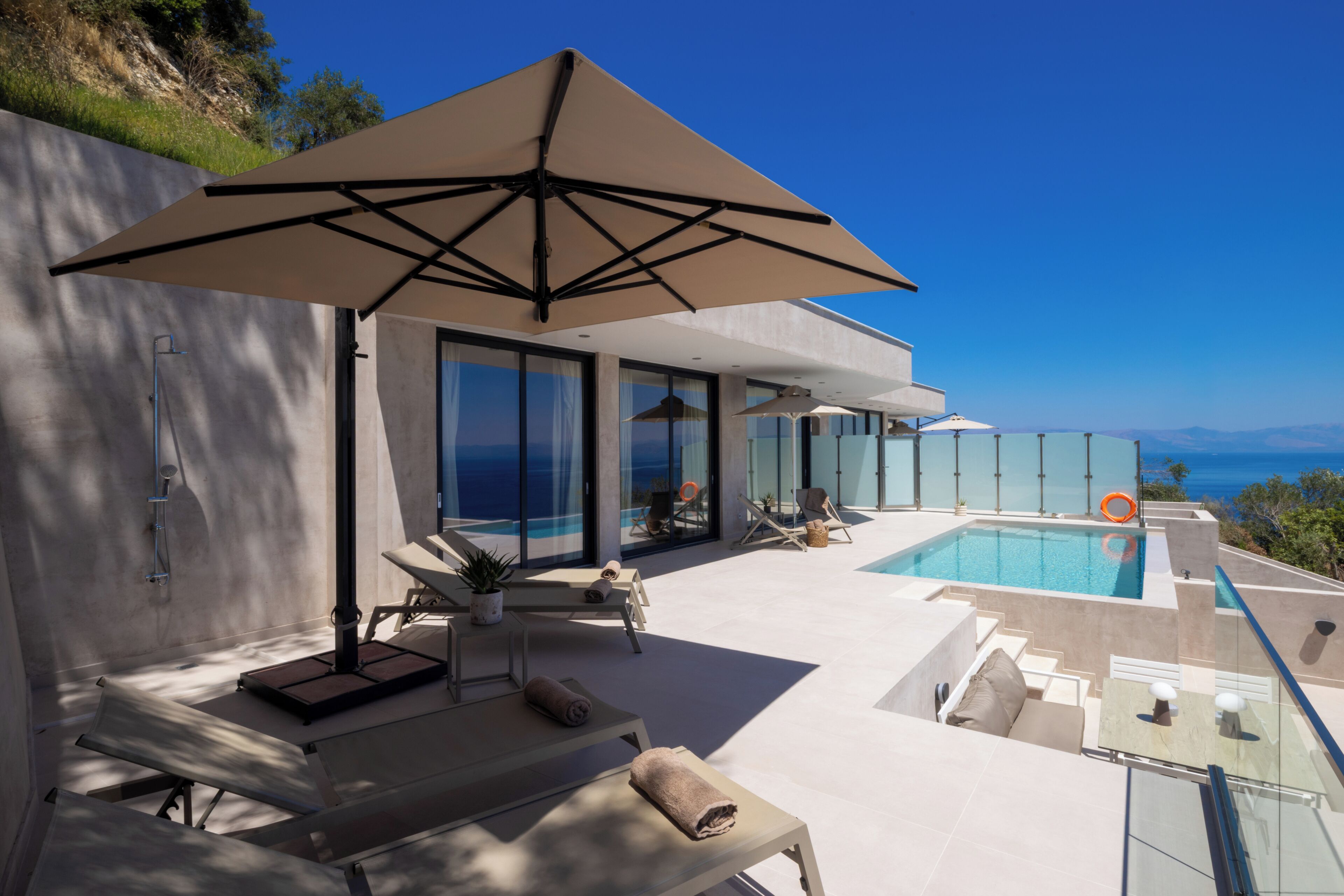 A modern outdoor pool area with lounge chairs and a large umbrella, overlooking a clear blue sky and a distant view.