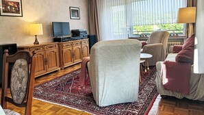 Un salon avec un meuble en bois, un téléviseur, un fauteuil à motifs et un tapis rouge à motifs.