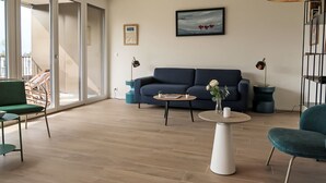 A modern living room with a blue sofa, a round coffee table, and a green armchair. There is a large window with a view of a balcony.