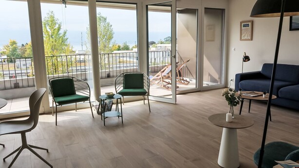 A modern living room with a large sliding glass door leading to a balcony, a blue sofa, a green chair, a small round table, and a floor lamp.