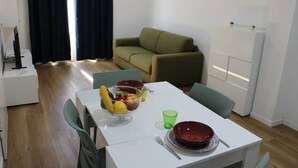 Un'area pranzo con un tavolo bianco apparecchiato per due persone, una ciotola di frutta e un bicchiere di vetro verde.