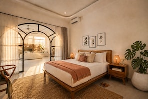A bedroom with a bed, bedside tables, a chair, a large plant, and a view into another room. - Villa Hamal by DVH Luxe Retreats 2BR (Canggu)