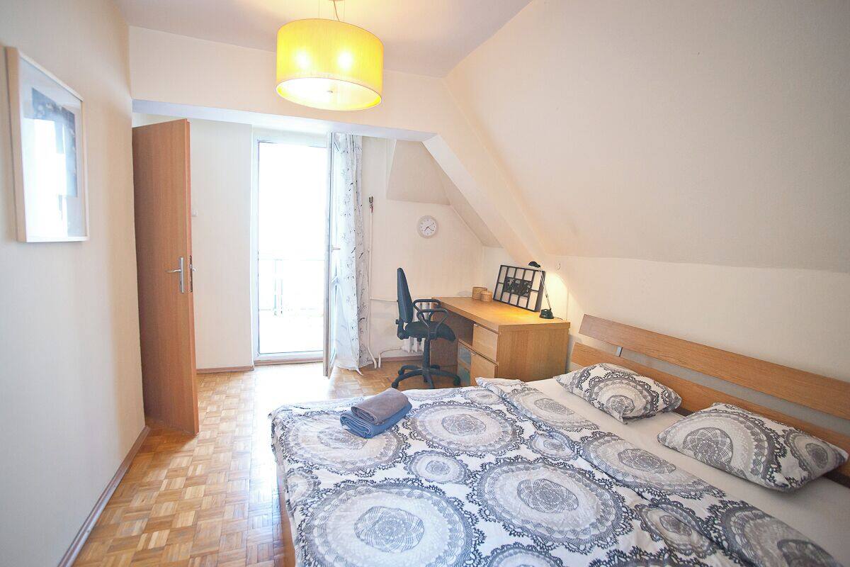 A bedroom with a bed, a desk, and a chair. There is a window with curtains, a door, and a clock on the wall.
