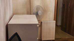 Una habitación con piso de madera, un radiador blanco, un ventilador de pie y una silla de oficina negra.