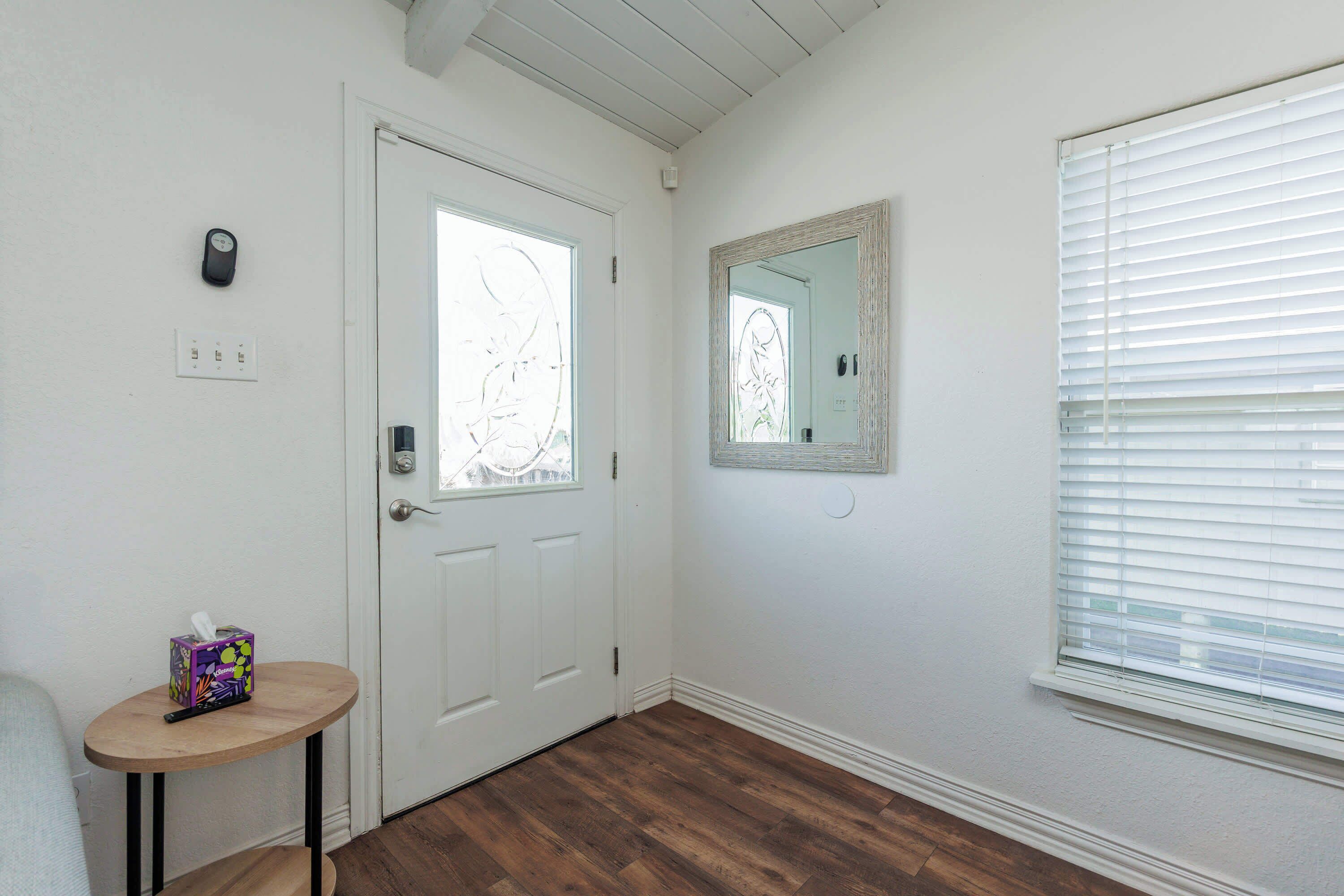 A hallway with a white door, a small table with a tissue box, a wall-mounted mirror, and a window with blinds.