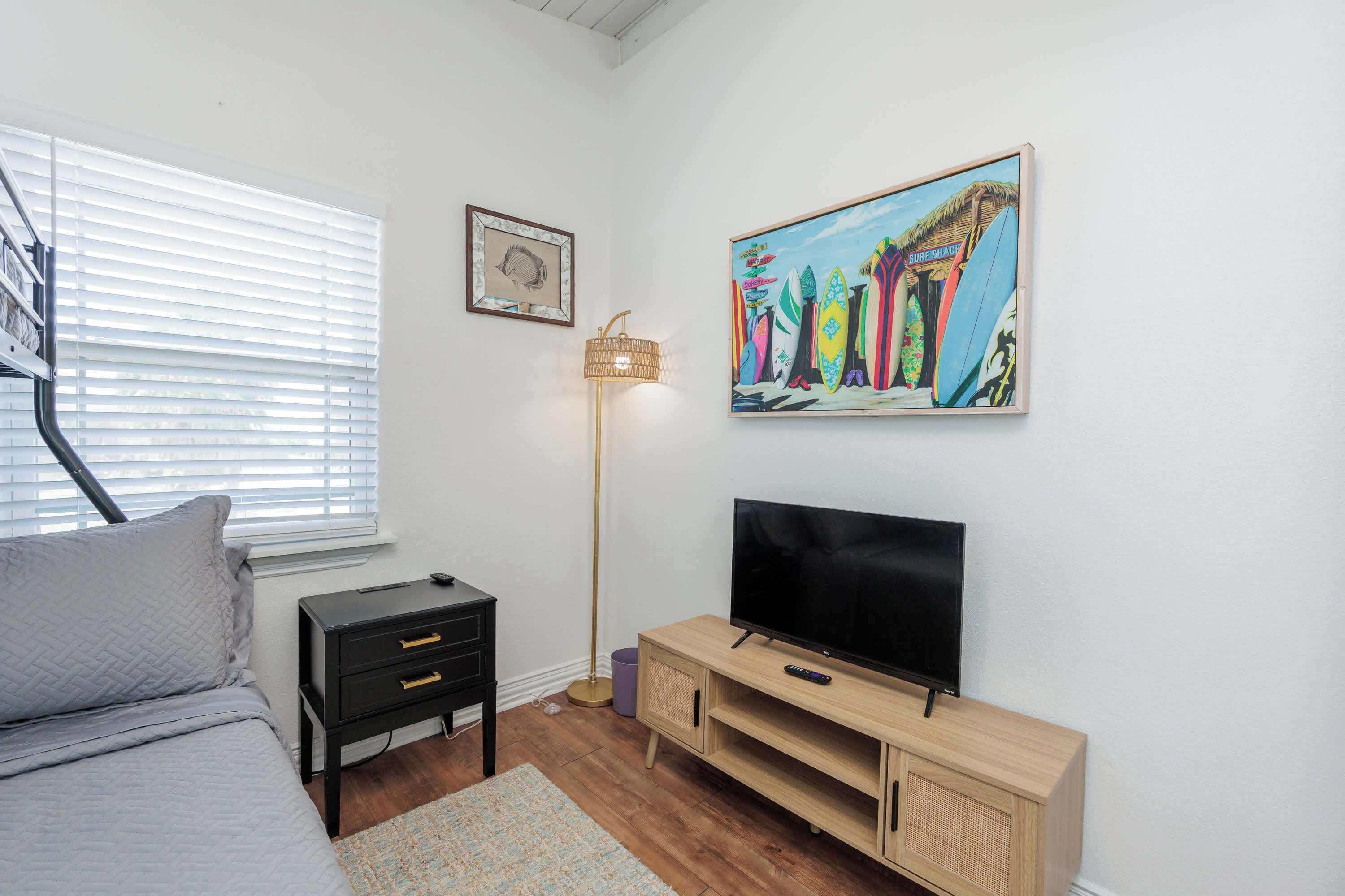 A bedroom with a bed, a TV on a wooden stand, a lamp, and a painting of surfboards.