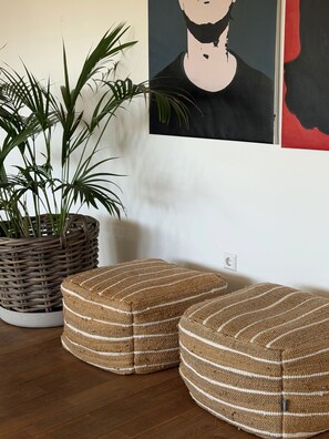 A room with a woven plant pot, two striped ottomans, a wall-mounted power outlet, and a partial view of a poster.