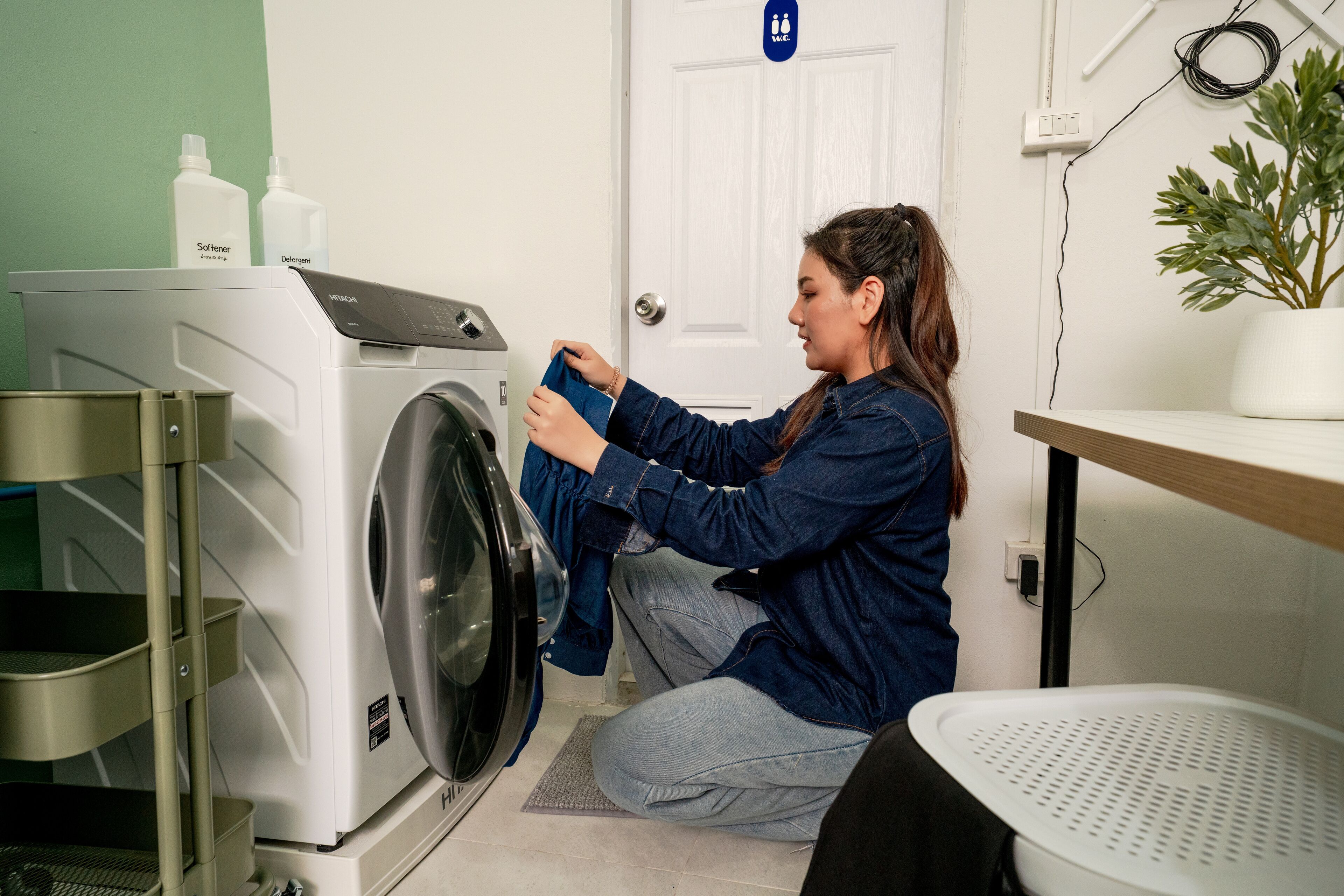 A person folding laundry in a room with a washing machine, a drying rack, and a potted plant.