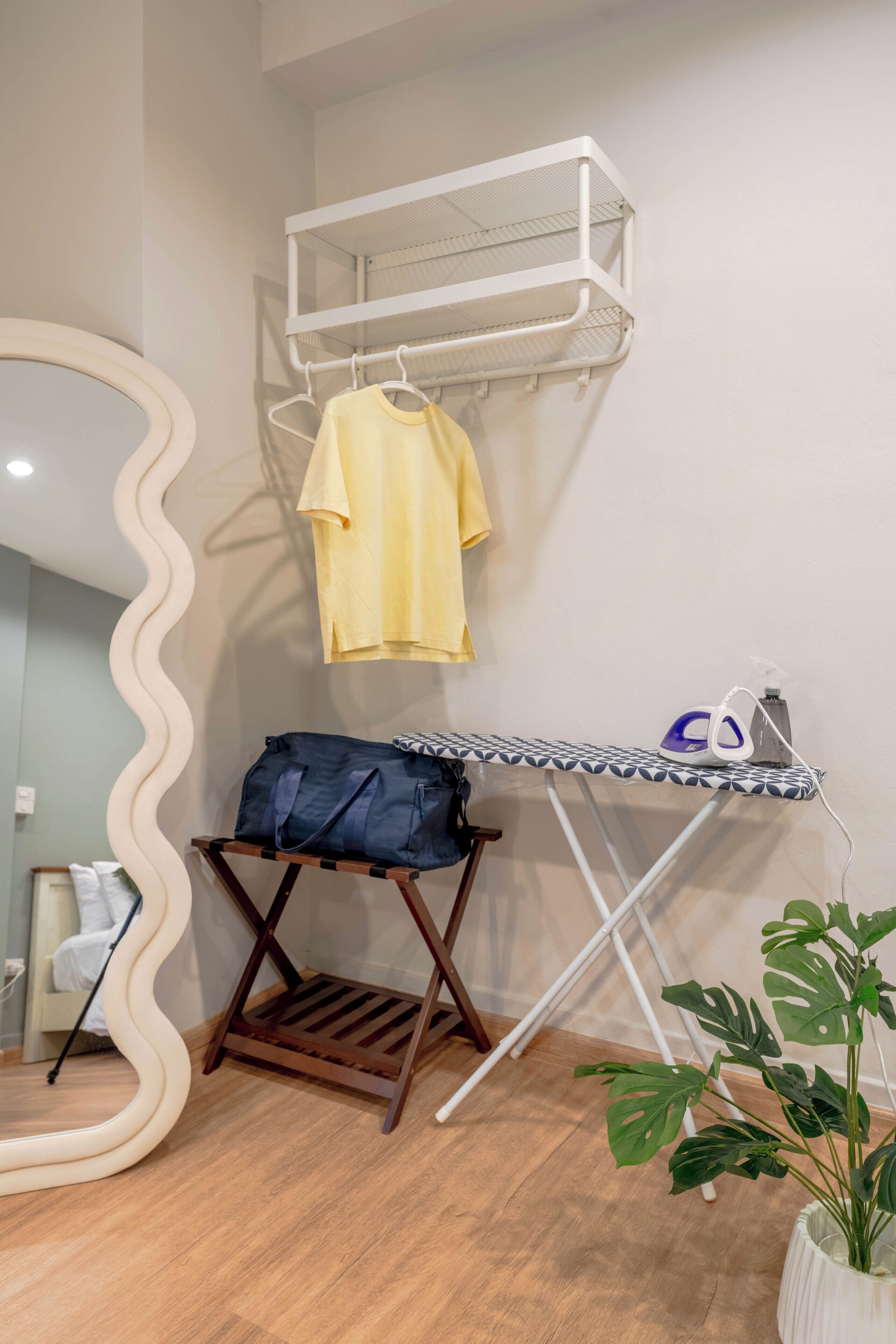 A neatly organized room with a wooden floor, a white mirror with a wavy frame, a hanging rack with clothes, an ironing board with an iron, and a potted plant.