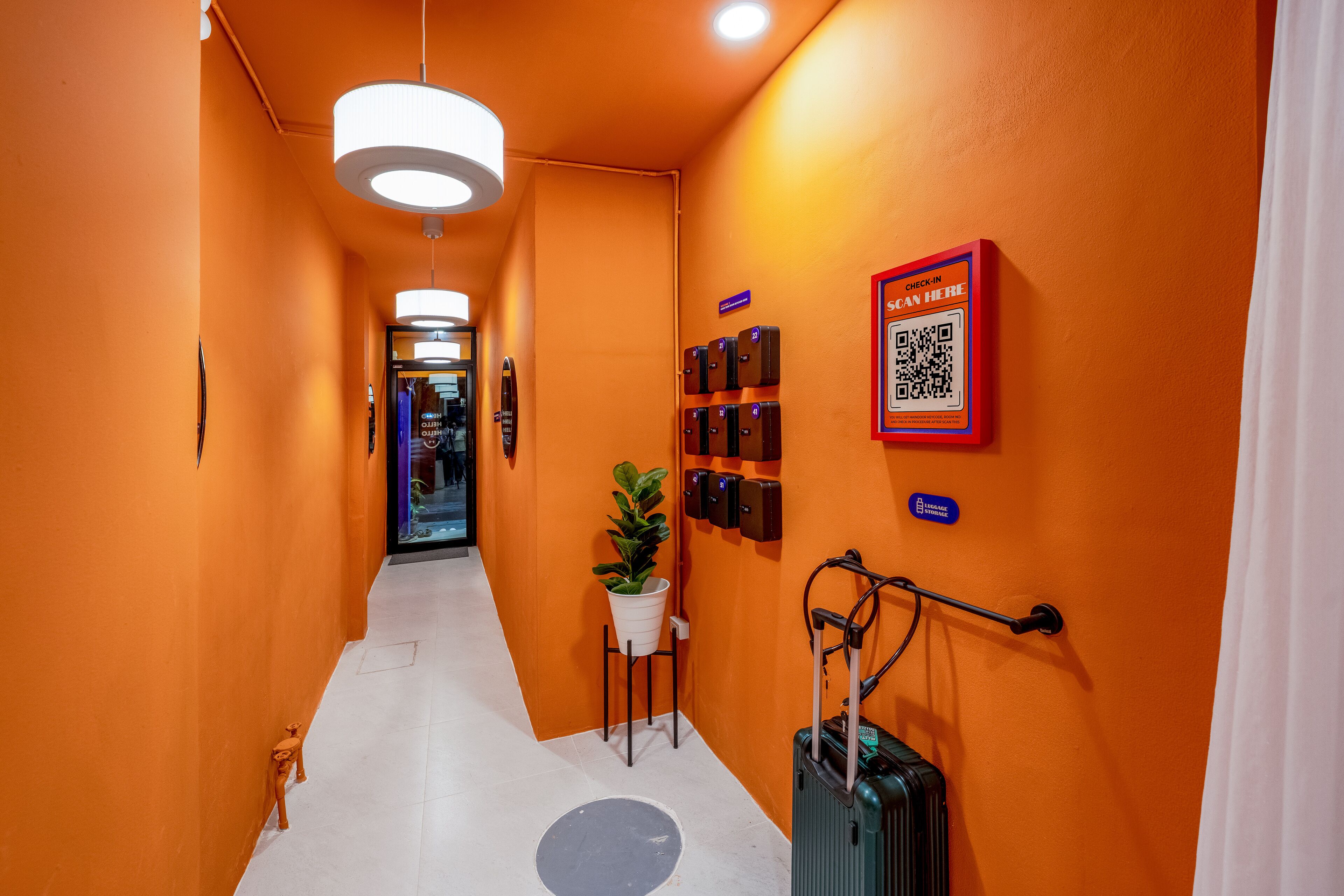 Pasillo de hotel con paredes naranja, un código QR para registrarse, espacio para guardar equipaje y una planta en maceta.