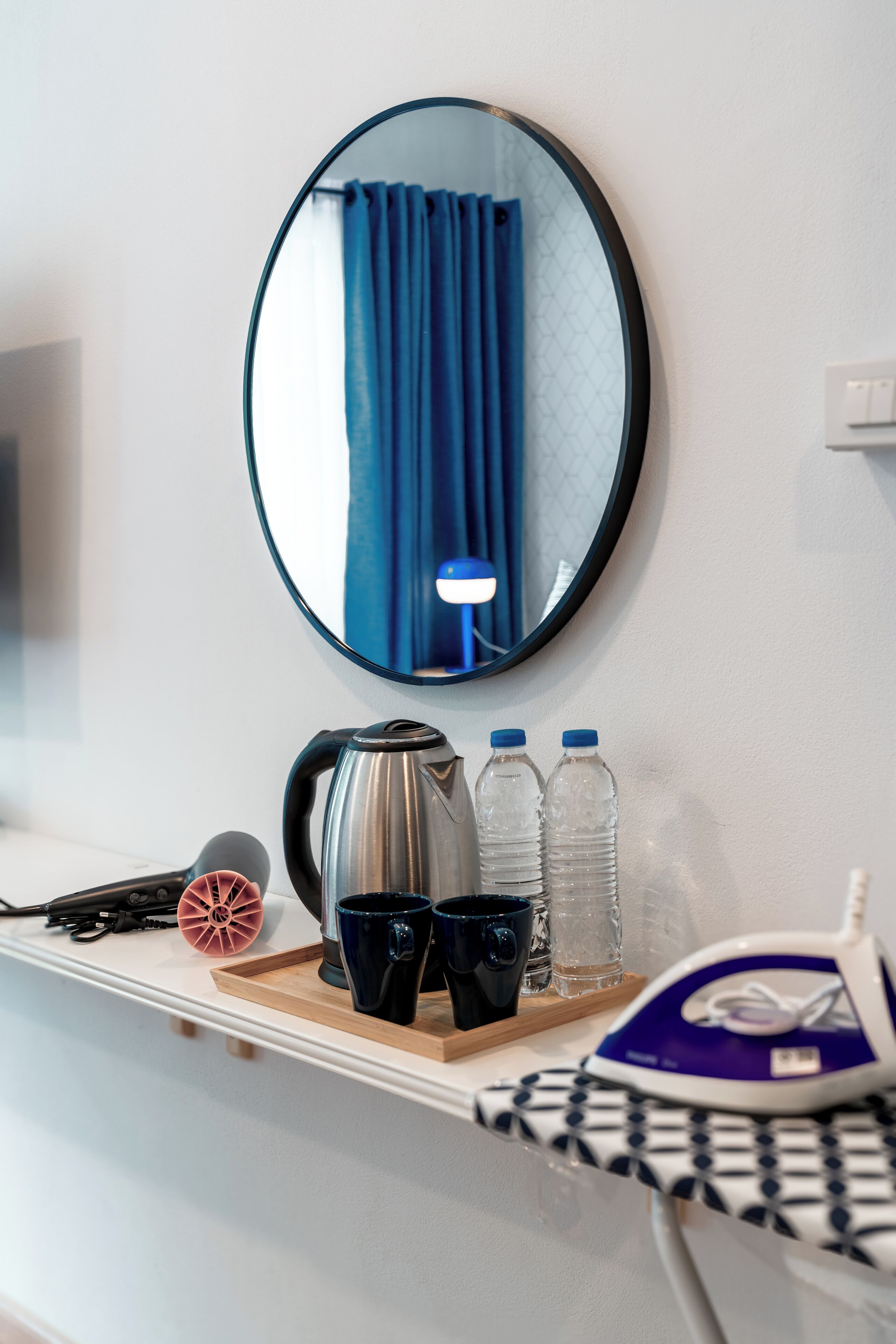 Hotelkamer met een ronde spiegel, een waterkoker, twee waterflessen, een koffiezetapparaat, een haardroger en een strijkplank.