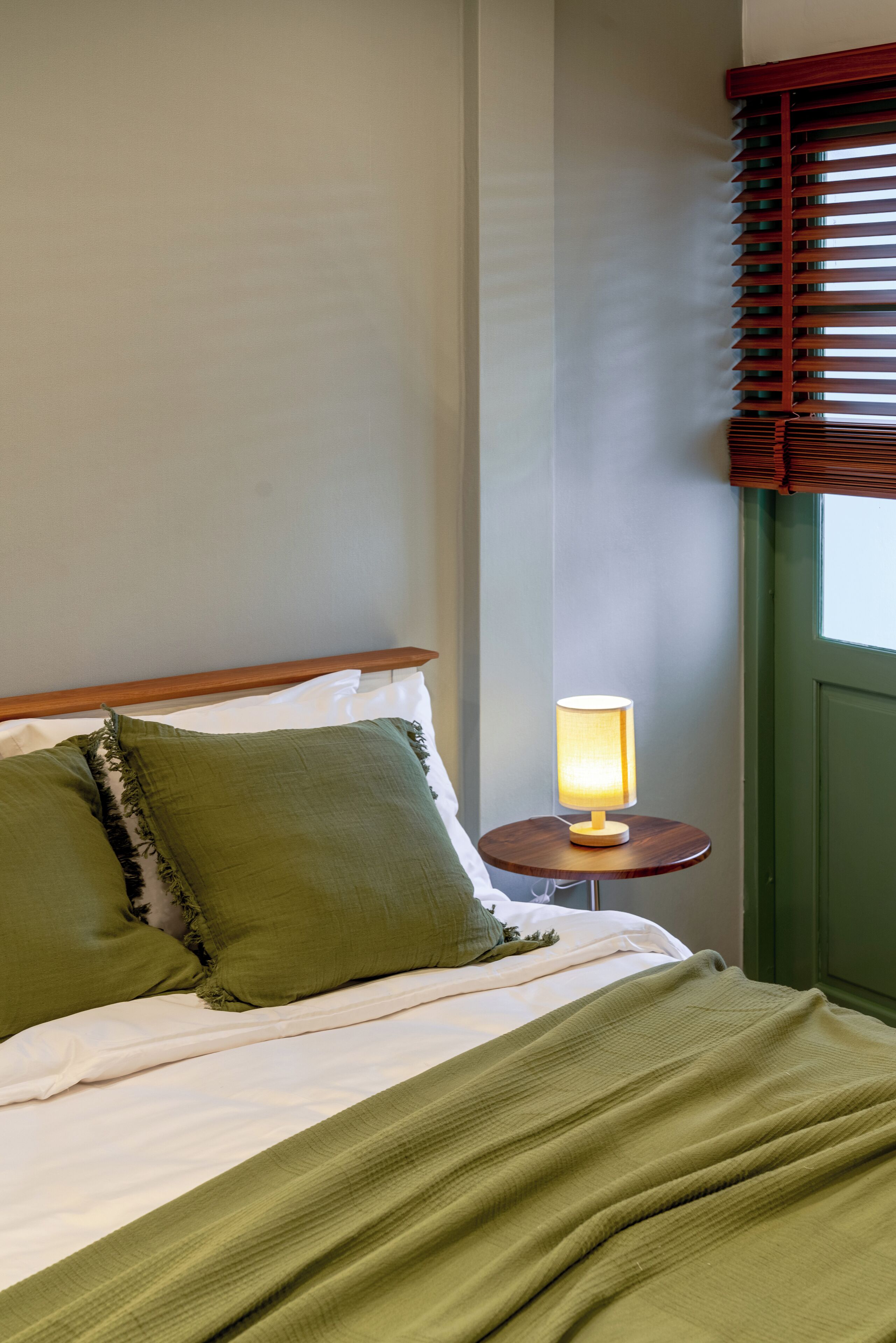 A neatly made bed with green pillows and a blanket, a bedside lamp, and a window with wooden blinds.