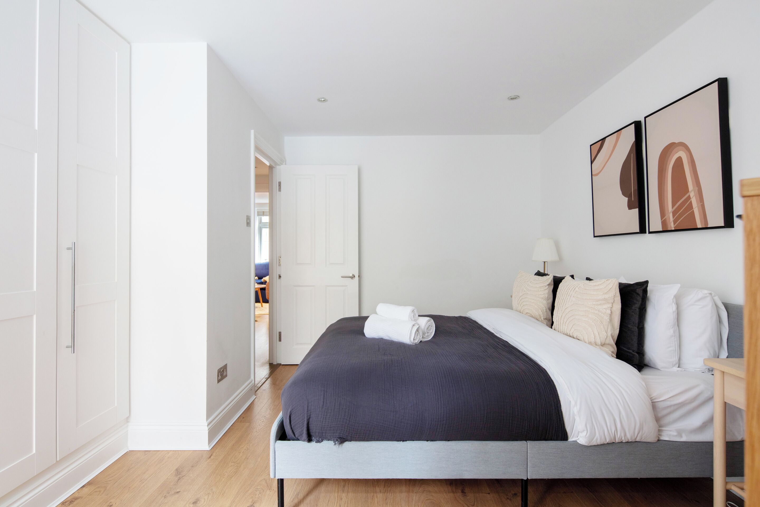 Ein modernes Schlafzimmer mit einem grauen Bett, weißen Wänden und Holzboden. Eine Tür führt in einen anderen Raum, und ein kleiner Tisch mit einer Lampe steht daneben.