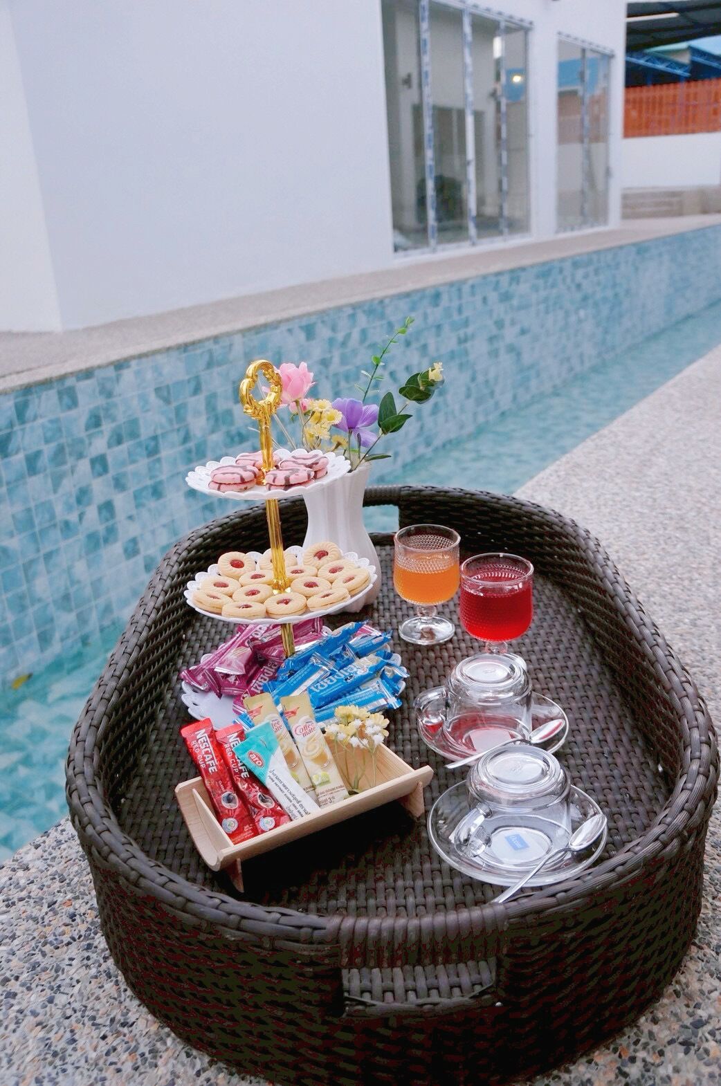 A wicker tray with snacks, drinks, and a flower arrangement by a poolside.