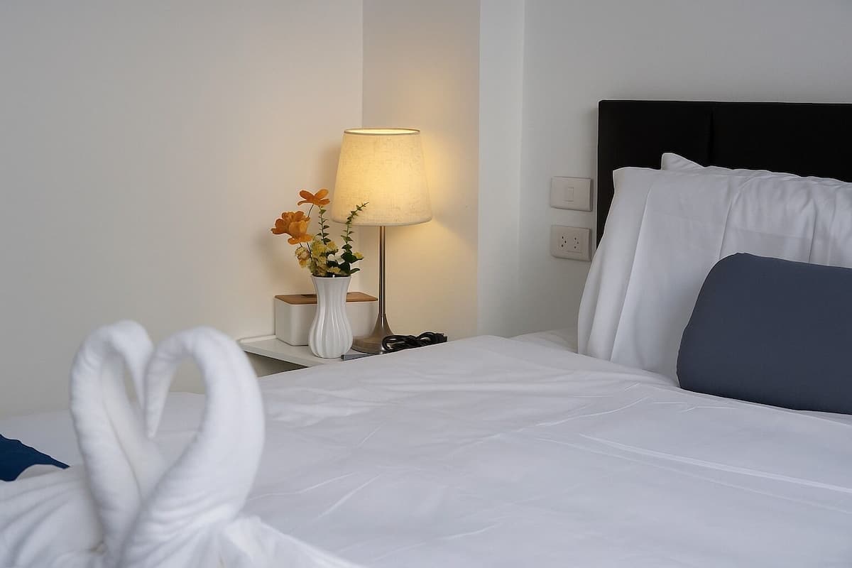 A neatly made bed with white linens, a bedside lamp, a vase with flowers, and a power outlet on the wall.