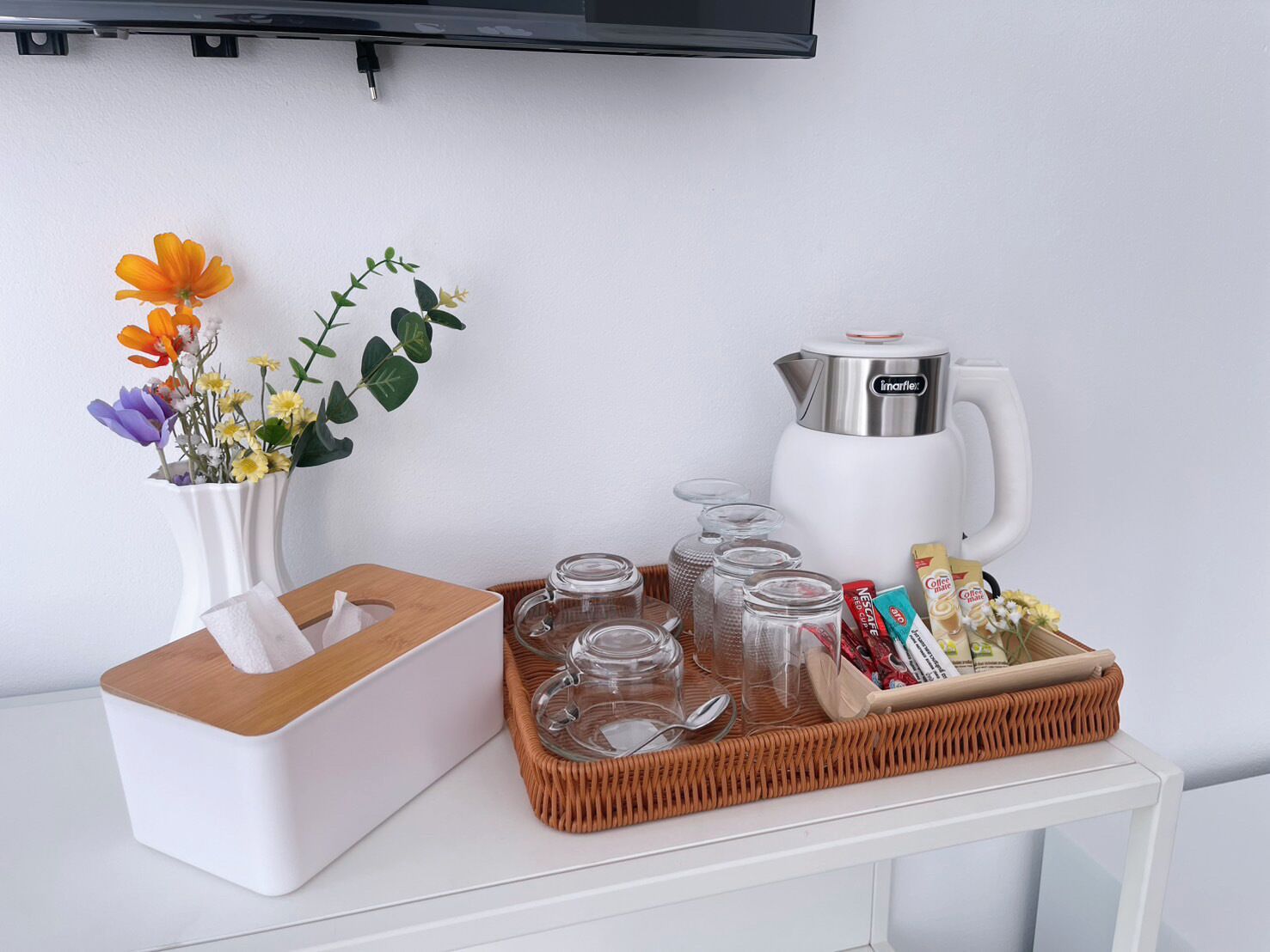 Ein ordentlich gedeckter Tisch mit Taschentuchbox, Blumen in einer Vase, einer Kaffeemaschine und einem Korb mit Gläsern und Snacks.