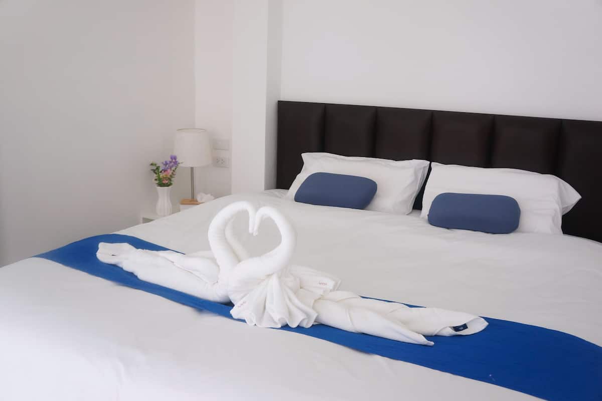 A neatly made bed with a blue bedspread and white pillows, a white vase with flowers, and a lamp on a bedside table.