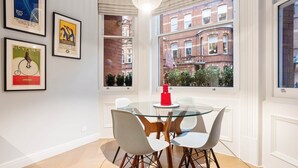 Un coin repas avec une table en verre et des chaises blanches, des affiches encadrées au mur et une vue sur un bâtiment en briques rouges, visible à travers de grandes fenêtres.