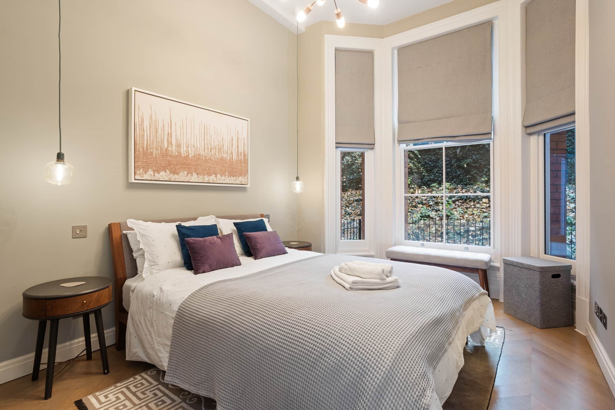 A bedroom with a large bed, a bedside table, a window with a view of greenery, and a painting on the wall.