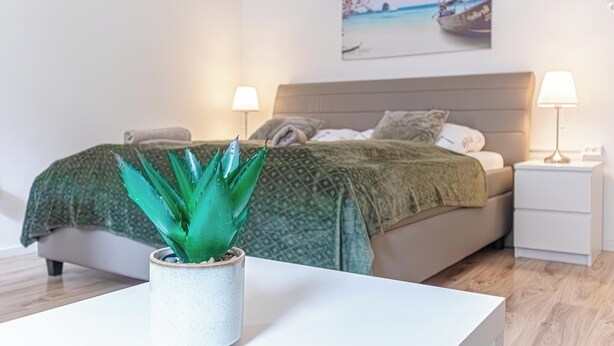 A bedroom with a bed, bedside lamps, a nightstand, a potted plant, and a painting on the wall.