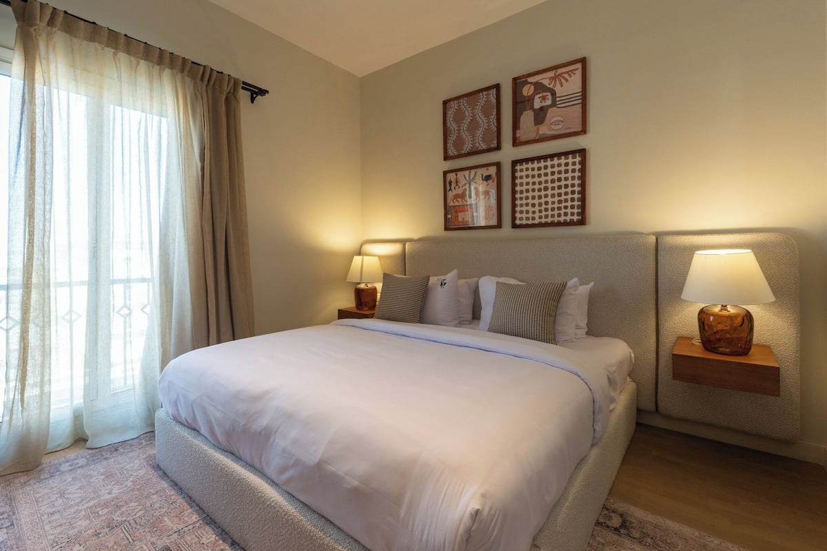 A hotel room with a large bed, two bedside tables with lamps, a window with curtains, and wall art.