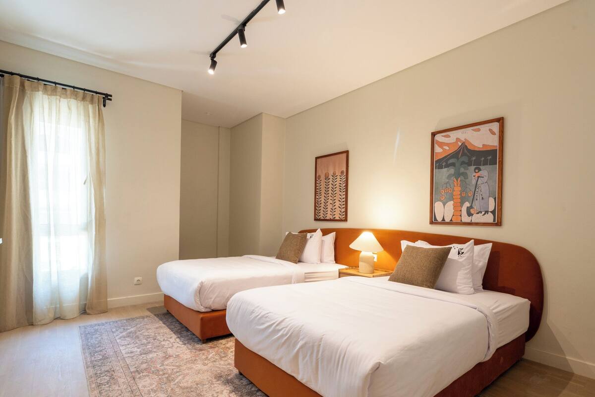 A double room with two beds, a large window with sheer curtains, a patterned rug, and two wall-mounted artworks.