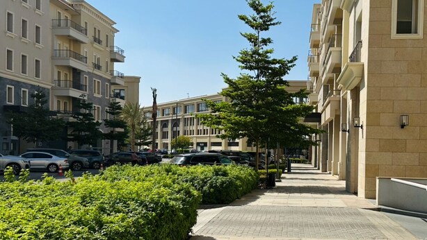 Ένα πολυώροφο κτίριο κατοικιών με μπαλκόνια, ένα πλακόστρωτο πεζοδρόμιο και σταθμευμένα αυτοκίνητα.
