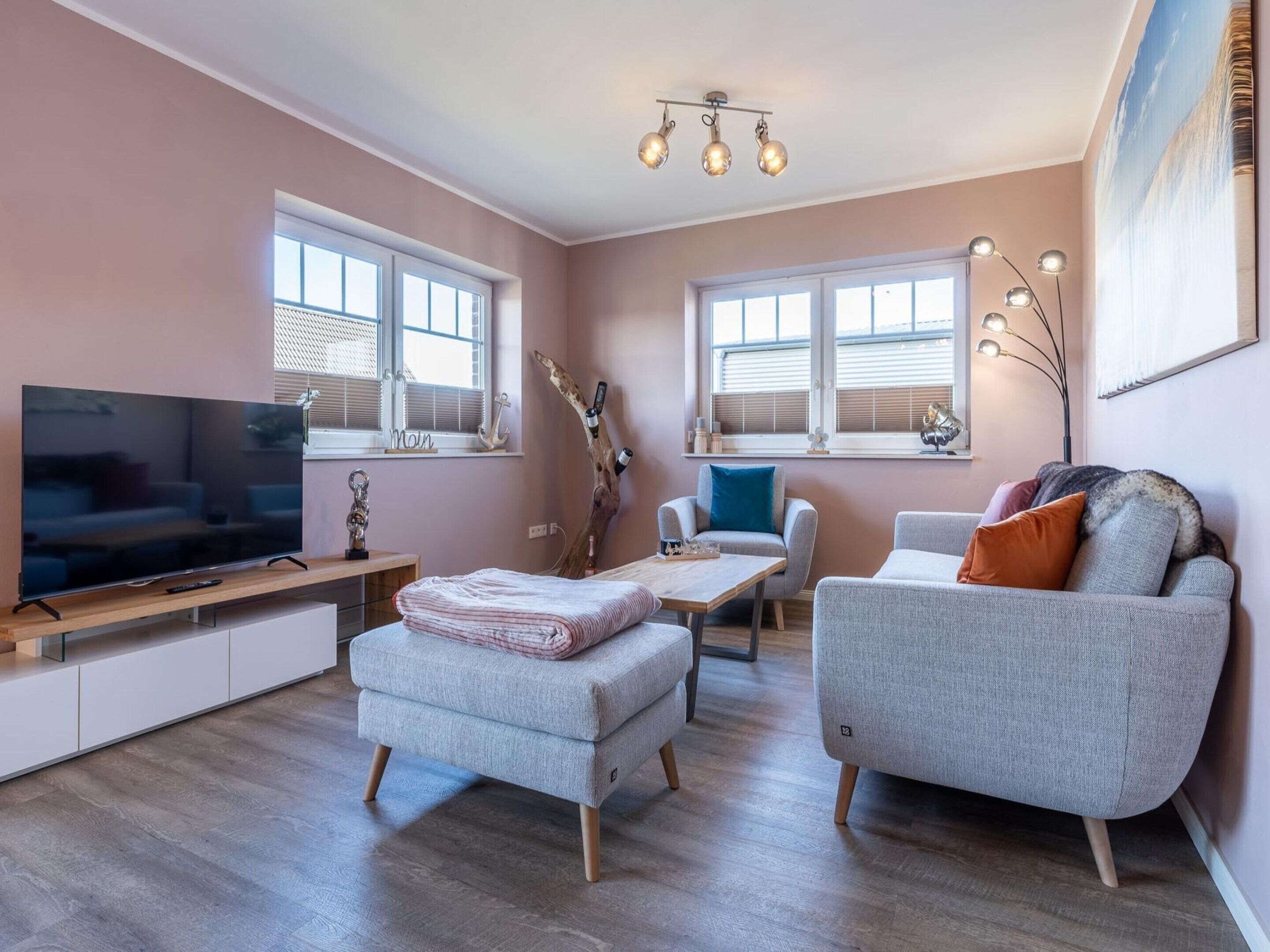 A modern living room with a flat-screen TV, a wooden coffee table, two grey sofas, and a large window with blinds.