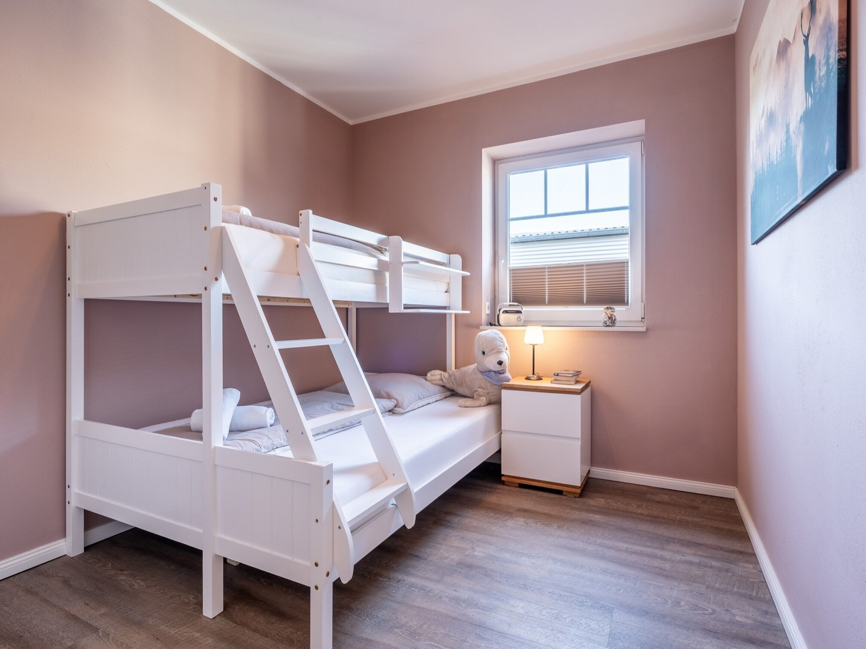 A bunk bed room with a ladder, a bedside table with a lamp, and a window with blinds.