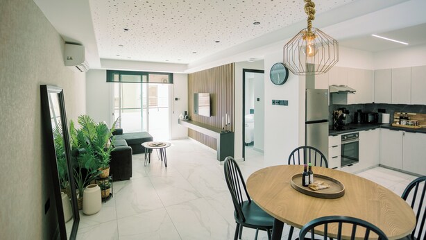 A modern kitchen with a central island, a dining area with a wooden table, and a living space with a sofa and plants. - Family-Friendly 2BR Apt at Mackenzie Area, Pool available, 10 Min Walk to Beach! (Larnaka)
