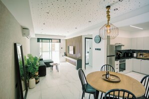 A modern kitchen with a central island, a dining area with a wooden table, and a living space with a sofa and plants.